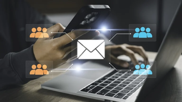 Person typing on a laptop and smartphone with a central inbox icon connected to orange and blue group icons, illustrating email distribution to groups.